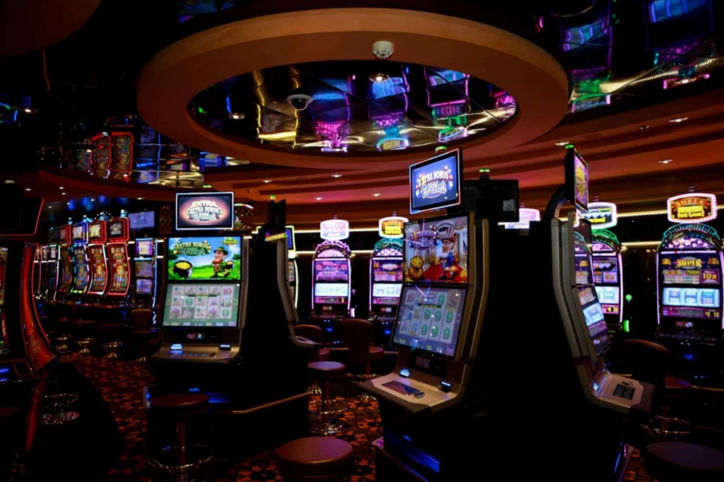 Salle de casino avec plusieurs rangées de machines à sous sous un plafond rond avec des néons colorés.